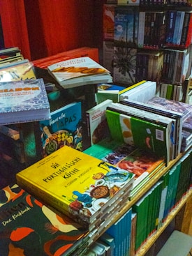 A cluttered and vibrant bookstand filled with a variety of cookbooks and novels. The books are stacked in different orientations, some are upright while others are laid flat. The covers display bright colors and images, particularly focusing on cuisines with titles in different languages, including Portuguese and German.