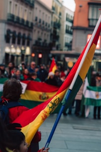 A crowd of people is gathered in a city street, holding colorful flags with red, yellow, and green colors. The architecture in the background features traditional European-style buildings, creating a lively urban atmosphere.