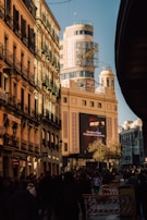 A bustling street scene with a tall building featuring a prominent Schweppes sign. The architecture is classic and illuminated by warm sunlight, casting shadows on the street below. Many people are walking, suggesting a lively urban atmosphere. A large advertisement screen displays a message in Spanish.