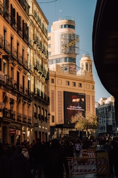A bustling street scene with a tall building featuring a prominent Schweppes sign. The architecture is classic and illuminated by warm sunlight, casting shadows on the street below. Many people are walking, suggesting a lively urban atmosphere. A large advertisement screen displays a message in Spanish.