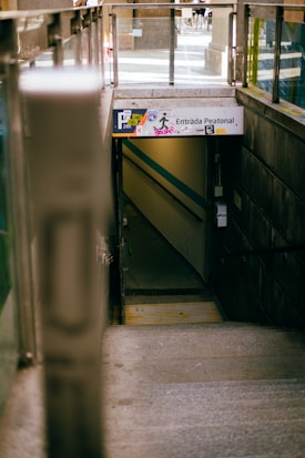 A concrete staircase descends into a narrow underground passageway labeled 'Entrada Peatonal' with graffiti on the sign. The passageway is dimly lit, and glass railings are on either side at the entrance from the street above.