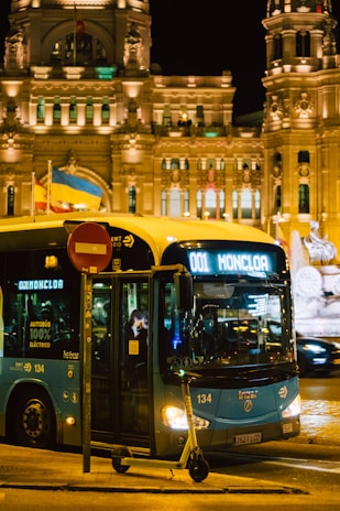 An illuminated electric bus labeled '001 Moncloa' is parked on a city street at night. The scene includes a grand, ornately lit building in the background, giving an urban and historic feel. A scooter is parked at the curb in front of the bus. Two flags, one blue and yellow, stand out on a pole near the bus.