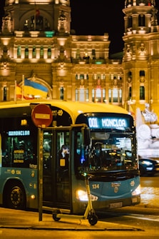 An illuminated electric bus labeled '001 Moncloa' is parked on a city street at night. The scene includes a grand, ornately lit building in the background, giving an urban and historic feel. A scooter is parked at the curb in front of the bus. Two flags, one blue and yellow, stand out on a pole near the bus.