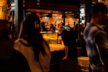 A bustling indoor market or food hall with people gathered around a food stall. The warm, ambient lighting highlights shelves filled with bottles and glasses hanging above. A person is leaning over a counter, possibly examining food items, while others move about, some appearing blurred.