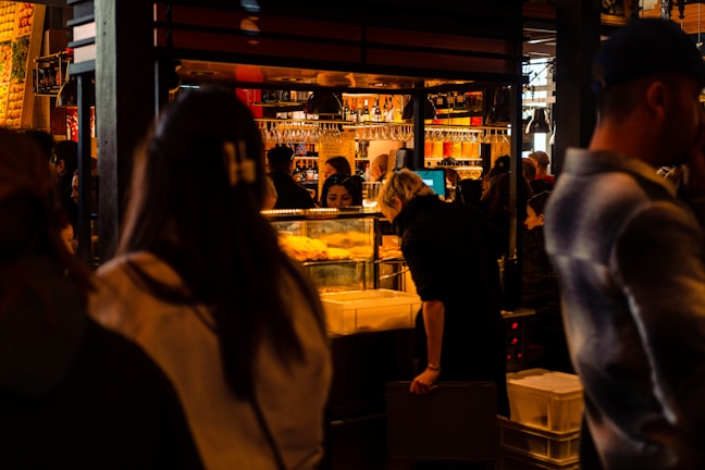 A bustling indoor market or food hall with people gathered around a food stall. The warm, ambient lighting highlights shelves filled with bottles and glasses hanging above. A person is leaning over a counter, possibly examining food items, while others move about, some appearing blurred.