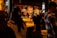 A bustling indoor market or food hall with people gathered around a food stall. The warm, ambient lighting highlights shelves filled with bottles and glasses hanging above. A person is leaning over a counter, possibly examining food items, while others move about, some appearing blurred.