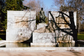 A stone monument featuring a central sculpture of a standing figure flanked by intricate carvings. Below, another sculpture depicts a seated figure. The monument includes two fountains that feed water into a shallow pool. Bare tree branches cast shadows on the structure, and there is grass and trees in the background.