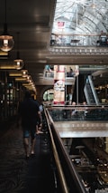 A multi-level shopping center with ornate architectural details and a high glass ceiling. Warm lighting from hanging lamps adds ambience, while people walk and shop on both levels. An escalator connects the floors, and banners advertise food and retail options.