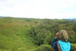 A person wearing a brown hood and a blue backpack is looking through binoculars towards a vast, lush green landscape with rolling hills and dense forest. The sky is overcast with some bright areas.