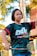 A person with short hair is seen holding a badminton racket. They are wearing a black t-shirt with colorful text and graphics, and they appear to be in an outdoor setting with trees and a building in the background.