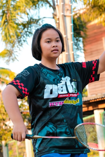 A person with short hair is seen holding a badminton racket. They are wearing a black t-shirt with colorful text and graphics, and they appear to be in an outdoor setting with trees and a building in the background.