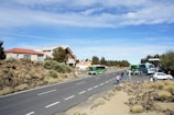 Several green buses and a few smaller vehicles are parked on the side of a wide road with people standing nearby. The area is surrounded by rocky terrain and sparse vegetation, with a few buildings with red and white roofs visible in the background. The sky is clear with scattered clouds, creating a bright and open atmosphere.