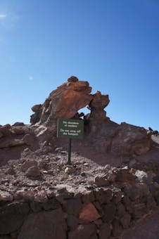 A rugged landscape with large rock formations under a clear blue sky. Prominently in the foreground is a sign that reads 'No abandone el sendero. Do not stray off the footpath,' indicating a designated walking path.