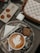 A marble-patterned tray holds a cappuccino with latte art in a ceramic cup, a neatly folded napkin, a knife, and a wooden stirrer. Next to the tray lies a small, square brownie. A smartphone with a decorative case featuring cartoon bear figures is placed nearby, along with a checkered designer purse and wireless earbud case.