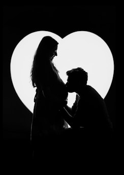 A silhouette of a pregnant woman and a man in front of a large, illuminated heart. The man is kneeling and appears to be kissing the woman's belly.