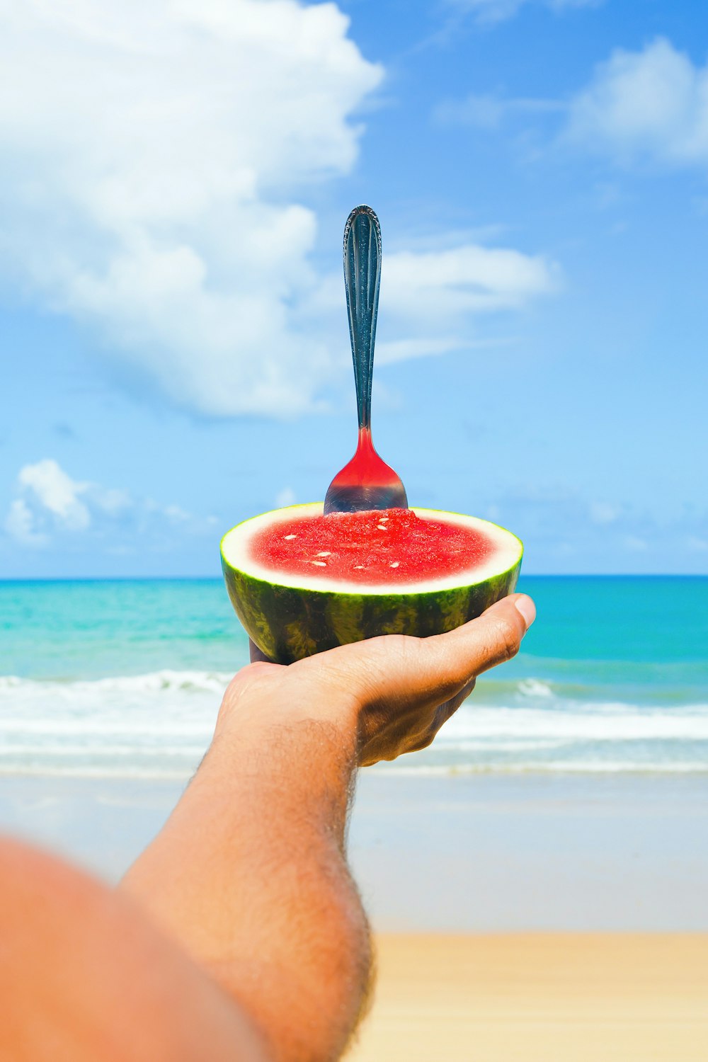 a hand holding a slice of watermelon on the beach