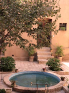 A serene courtyard features a circular blue fountain surrounded by potted plants and lush green foliage. The courtyard is paved with light-colored bricks, and steps lead up to a wooden door on the earthy-toned building wall. Sunshine filters through the leaves, creating a warm and inviting atmosphere.
