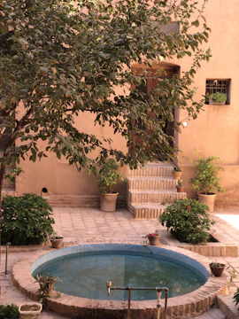 A serene courtyard features a circular blue fountain surrounded by potted plants and lush green foliage. The courtyard is paved with light-colored bricks, and steps lead up to a wooden door on the earthy-toned building wall. Sunshine filters through the leaves, creating a warm and inviting atmosphere.