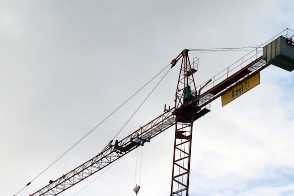 A large construction crane is captured against a cloudy sky. The crane is built with a lattice framework and features a prominent yellow sign with the word 'Azvi' on it.