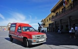 A red vehicle is parked on a wide paved area near a building with a long veranda. A police officer with a helmet stands nearby. Several people walk in the background under a clear blue sky.