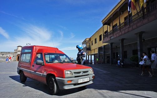 A red vehicle is parked on a wide paved area near a building with a long veranda. A police officer with a helmet stands nearby. Several people walk in the background under a clear blue sky.