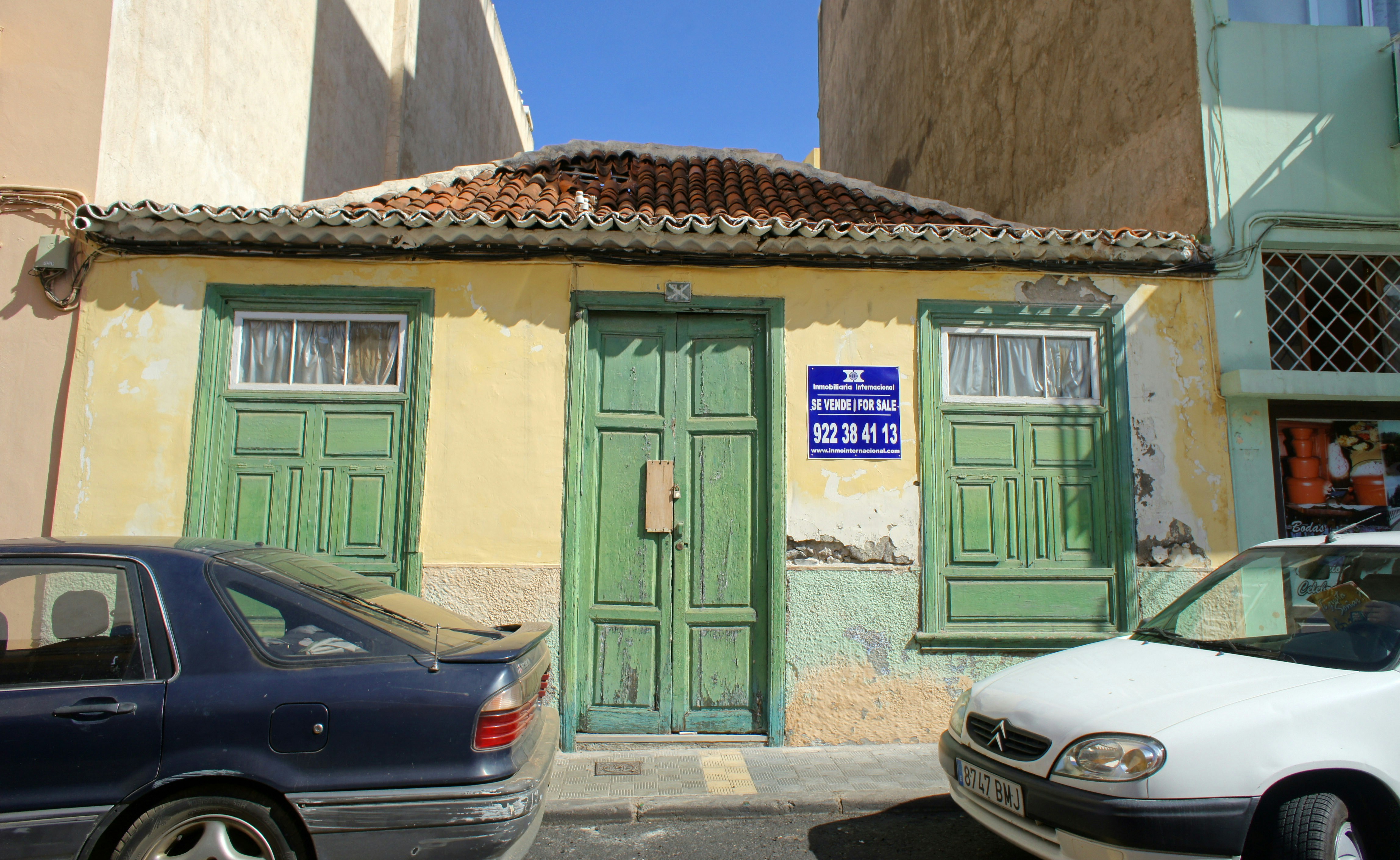An old, weathered building with green doors and windows, featuring a textured yellow and faded green facade. The roof is made of terracotta tiles, and the building has a 'For Sale' sign with contact information written in Spanish and English. Two cars, a dark blue one and a white one, are parked in front. The building seems situated in a bright, sunny area.