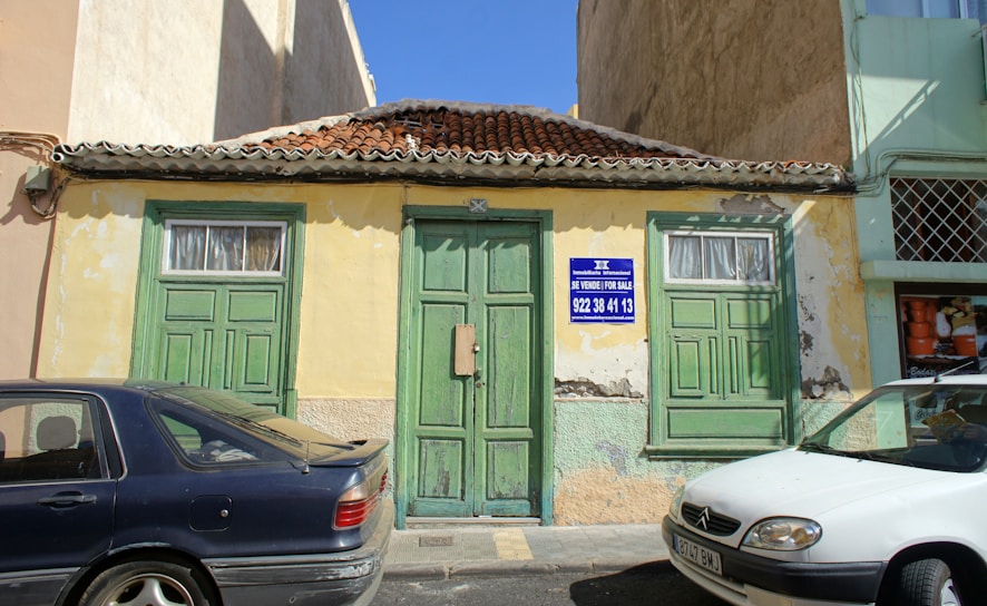 An old, weathered building with green doors and windows, featuring a textured yellow and faded green facade. The roof is made of terracotta tiles, and the building has a 'For Sale' sign with contact information written in Spanish and English. Two cars, a dark blue one and a white one, are parked in front. The building seems situated in a bright, sunny area.
