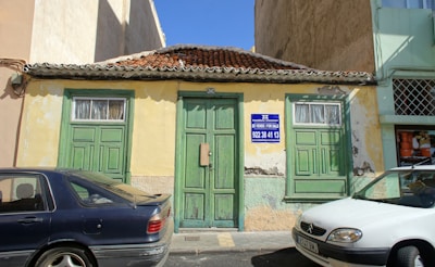 An old, weathered building with green doors and windows, featuring a textured yellow and faded green facade. The roof is made of terracotta tiles, and the building has a 'For Sale' sign with contact information written in Spanish and English. Two cars, a dark blue one and a white one, are parked in front. The building seems situated in a bright, sunny area.
