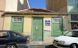 An old, weathered building with green doors and windows, featuring a textured yellow and faded green facade. The roof is made of terracotta tiles, and the building has a 'For Sale' sign with contact information written in Spanish and English. Two cars, a dark blue one and a white one, are parked in front. The building seems situated in a bright, sunny area.