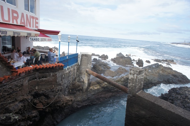 A seaside restaurant with people dining under a red awning. The building is situated on rugged rocks by the ocean, with waves crashing in the background. A blue railing separates the dining area from the rocks and ocean. The sky is partly cloudy, creating a serene coastal atmosphere.