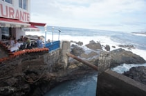 A seaside restaurant with people dining under a red awning. The building is situated on rugged rocks by the ocean, with waves crashing in the background. A blue railing separates the dining area from the rocks and ocean. The sky is partly cloudy, creating a serene coastal atmosphere.