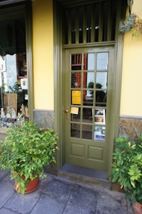 A storefront entrance features a green door with glass panes, surrounded by a yellow wall. Two potted plants are placed symmetrically on either side of the door. A small yellow sign with black text is affixed to the door. Through the glass, various items and some greenery are visible inside the shop.