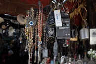 An assortment of vintage objects including old cameras, a Polaroid camera, and a cassette recorder are hanging alongside beaded strings and a large decorative evil eye amulet. The background is filled with various eclectic items such as pottery and black-and-white photographs, hinting at an antique or flea market setting.
