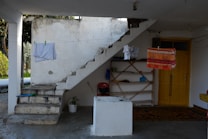 An outdoor area features a weathered set of concrete stairs with laundry hanging on a line above. A yellow door is on the right, next to a set of shelves with various items including a blue dustpan and a red watering can. A patterned rug covers part of the floor, and a small potted plant is placed near the stairs.