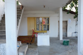 A simple outdoor space with concrete steps leading up, a wooden door, and a window with a white curtain. There is a red plastic chair next to the door and a decorative rug on the ground. A small line holding laundry is visible, along with a green plastic crate on the ground. The area is partially covered by a roof with an exposed beam and surrounded by white walls.