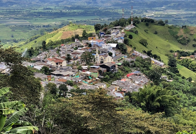 A small town is nestled among lush green hills and mountainous terrain. The town features a mixture of colorful buildings with a prominent church in the center. It is surrounded by dense vegetation, farmland, and winding roads that connect the settlement to the broader rural landscape.