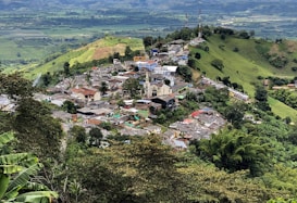 A small town is nestled among lush green hills and mountainous terrain. The town features a mixture of colorful buildings with a prominent church in the center. It is surrounded by dense vegetation, farmland, and winding roads that connect the settlement to the broader rural landscape.