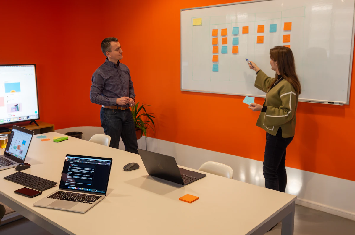 In a meeting room, a woman points at columns of sticky notes on a whiteboard while a colleague stands back and listens