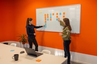 Two women are standing in a brightly colored office room with an orange wall. They are discussing or arranging colored sticky notes on a whiteboard. There is a table in the foreground with a laptop, a coffee mug, a mouse, and more sticky notes. A plant is placed near the wall.