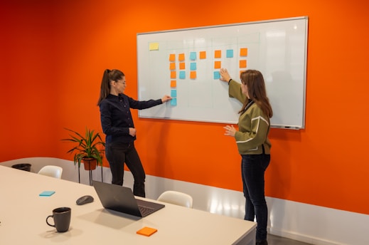Two women are standing in a brightly colored office room with an orange wall. They are discussing or arranging colored sticky notes on a whiteboard. There is a table in the foreground with a laptop, a coffee mug, a mouse, and more sticky notes. A plant is placed near the wall.