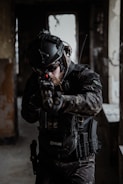A person wearing tactical gear and a helmet aims a firearm in a dimly lit, possibly abandoned building. The gear includes protective padding, gloves, and a radio communication device. The lighting is low, creating a tense and focused atmosphere.