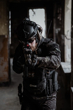 A person wearing tactical gear and a helmet aims a firearm in a dimly lit, possibly abandoned building. The gear includes protective padding, gloves, and a radio communication device. The lighting is low, creating a tense and focused atmosphere.