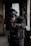 A person wearing tactical gear and a helmet aims a firearm in a dimly lit, possibly abandoned building. The gear includes protective padding, gloves, and a radio communication device. The lighting is low, creating a tense and focused atmosphere.