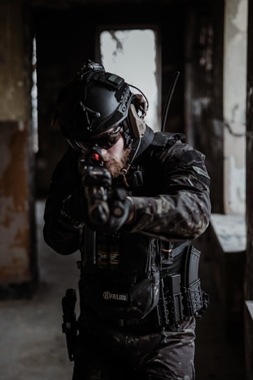 A person wearing tactical gear and a helmet aims a firearm in a dimly lit, possibly abandoned building. The gear includes protective padding, gloves, and a radio communication device. The lighting is low, creating a tense and focused atmosphere.