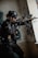 A tactical soldier dressed in full gear and helmet is aiming a rifle while standing indoors against a wall with graffiti. The soldier wears sunglasses and has various tactical equipment attached to their vest. The setting appears to be an urban, possibly abandoned structure.