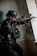 A tactical soldier dressed in full gear and helmet is aiming a rifle while standing indoors against a wall with graffiti. The soldier wears sunglasses and has various tactical equipment attached to their vest. The setting appears to be an urban, possibly abandoned structure.