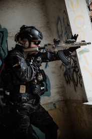 A tactical soldier dressed in full gear and helmet is aiming a rifle while standing indoors against a wall with graffiti. The soldier wears sunglasses and has various tactical equipment attached to their vest. The setting appears to be an urban, possibly abandoned structure.