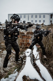 Two individuals in tactical gear are positioned outdoors with rifles, taking cover behind snow-covered logs. The scene appears to be in an urban setting, with an industrial-looking building in the background. They wear helmets and camouflage uniforms.