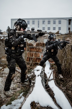Two individuals in tactical gear are positioned outdoors with rifles, taking cover behind snow-covered logs. The scene appears to be in an urban setting, with an industrial-looking building in the background. They wear helmets and camouflage uniforms.