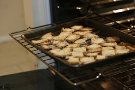 A baking tray filled with golden brown crackers is placed inside an oven. The crackers have a light dusting of what seems to be salt or cheese on their surface.